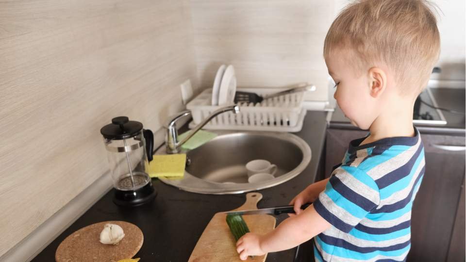Un jeune enfant coupe des légumes de manière autonome sur une planche à découper dans la cuisine.